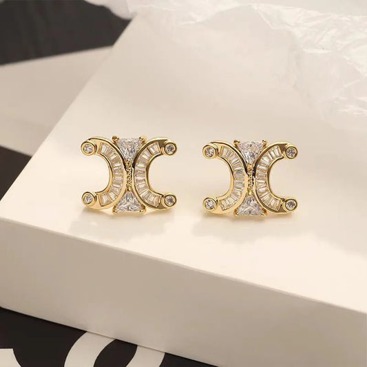 Gold-tone double-crescent stud earrings with clear baguette and round stones on a white box.