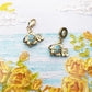 Two gold-tone fish dangle charms with blue accents, one with lobster clasp and one with bead bail on painted background