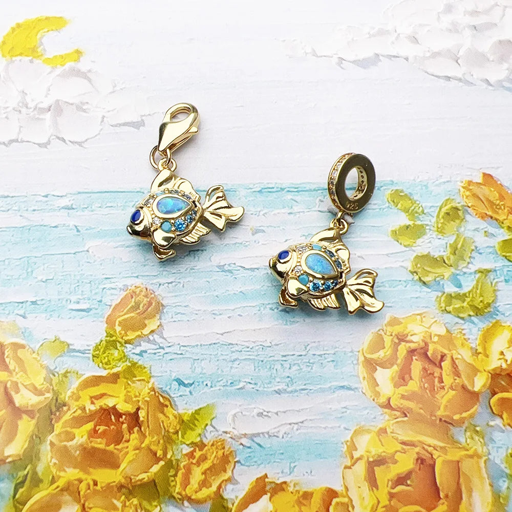 Two gold-tone fish dangle charms with blue accents, one with lobster clasp and one with bead bail on painted background