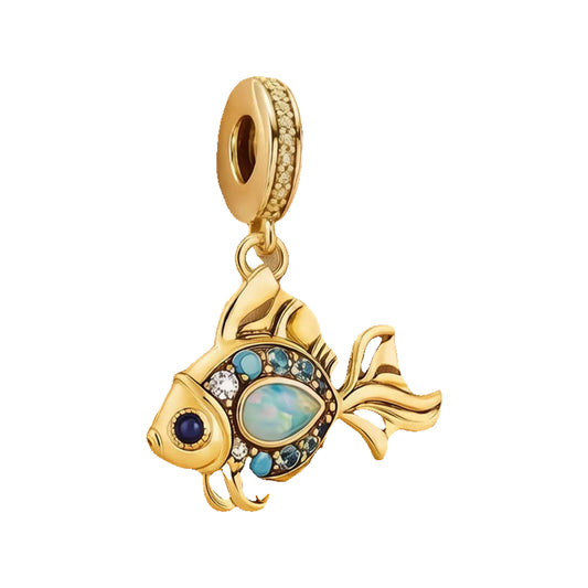 Gold-tone fish charm resting on satin fabric showing opal-look center, blue stones, and pavé bail