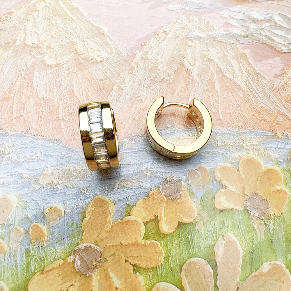 Two gold-tone huggie hoops, one showing the inner clasp and one the channel-set white stones on a floral painted background